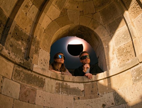 Une femme et un homme portant des lunettes de protection observent une éclipse solaire depuis une coupole historique en pierre, éclairée par la lumière chaleureuse de la fin de journée.