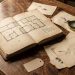 Nature morte Belle Époque avec un carnet d’enquête ouvert, un plan de chambre sans annotations, des feuilles anciennes, une loupe en laiton et de petites étiquettes vierges sur une table en bois.